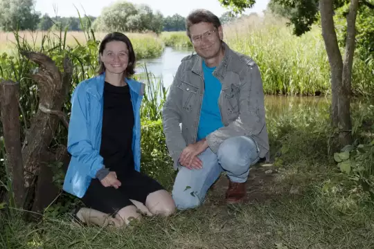 Zwei Personen knieend vor der Örtze und einer sommerlichen Wiesenlandschaft